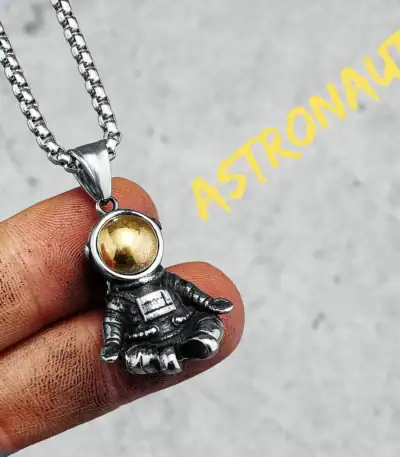 Close-up of a silver astronaut pendant necklace with a gold sphere helmet, held between fingers against a neutral background.