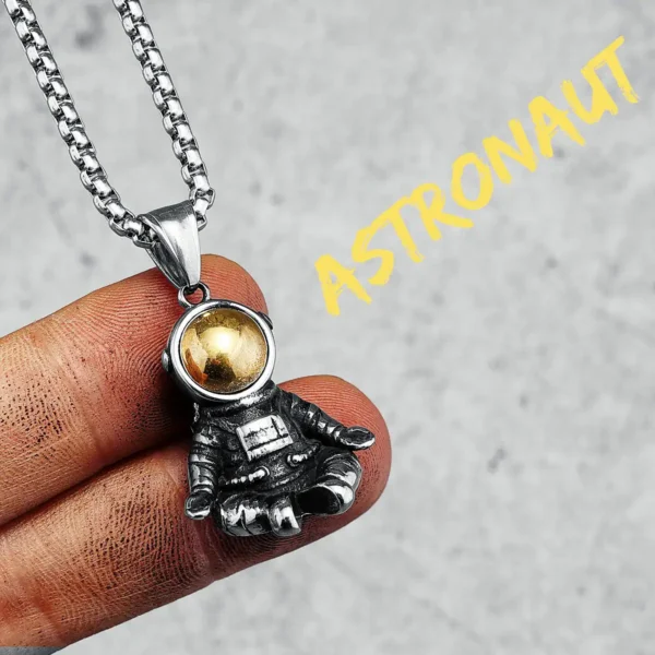 Close-up of a silver astronaut pendant necklace with a gold sphere helmet, held between fingers against a neutral background.