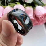 Hand holding a cosmic-themed resin ring with galaxy design, black band, and glitter accents, against a background of pink roses.