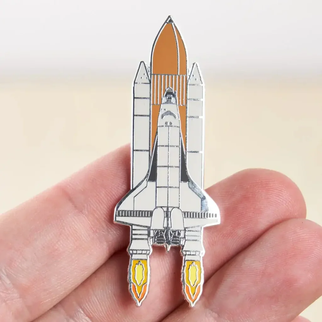 Small space shuttle pin with detailed design, held between fingers, perfect for science enthusiasts and collectors.