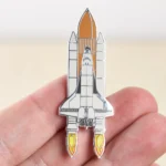Small space shuttle pin with detailed design, held between fingers, perfect for science enthusiasts and collectors.