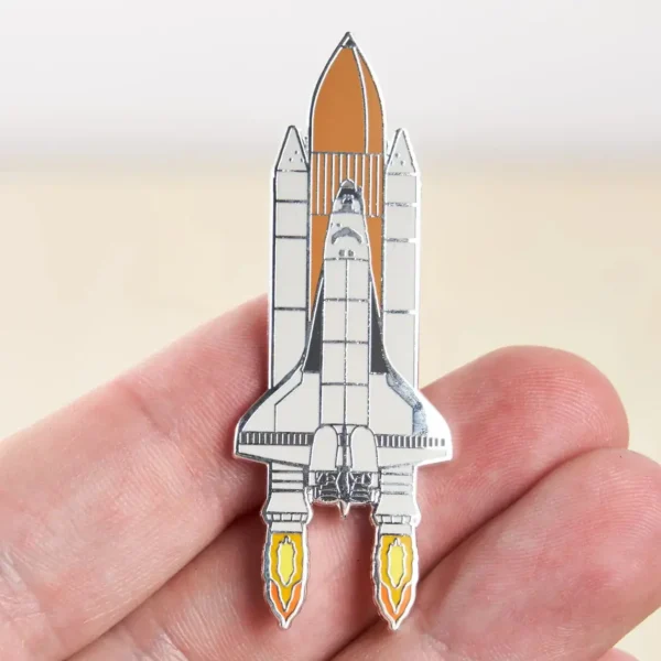 Small space shuttle pin with detailed design, held between fingers, perfect for science enthusiasts and collectors.