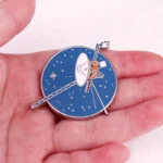 Enamel pin featuring a satellite spacecraft with solar panels and antenna, designed in blue, white, and orange colors, perfect for space enthusiasts.