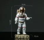 Astronaut figurine with a gold helmet, white suit with red accents, standing on a textured rock base, measuring approximately 22cm tall, 12cm wide, and 10cm deep.