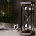Stylish glass hourglass timer with black sand, featuring a wooden and metal frame, ideal for educational and decorative purposes.
