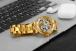Elegant gold skeleton watch with visible mechanical movement resting on a laptop keyboard, with wireless earbuds nearby, showcasing a blend of technology and luxury accessories.
