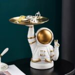 Astronaut figurine with a gold helmet holding a tray filled with science gadgets and accessories, set against a dark background.