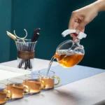 A hand is pouring hot water from a glass teapot with a built-in infuser into three transparent glass cups, showcasing a science-inspired tea brewing device.