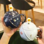 Two space-themed teapots with celestial designs, one dark blue and one white, featuring planets, stars, and moons, perfect for space lovers and science decor.