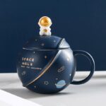 Dark blue ceramic mug featuring a space-themed design with stars, planets, and a gold astronaut figure on the lid, perfect for science and space enthusiasts.