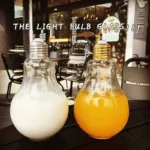 Creative light bulb-shaped glasses, one white and one yellow, displayed outdoors with a cafe background, showcasing unique drinkware design.