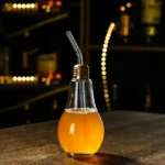 Creative light bulb-shaped glass filled with orange juice, featuring a metal straw, on a wooden surface with blurred warm lighting in the background.