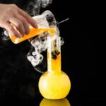 A science experiment showing a hand pouring orange liquid into a beaker with smoke and a test tube, set against a black background.