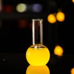 Bright yellow liquid in a laboratory flask with a long neck, illuminated against a dark background with bokeh lights.