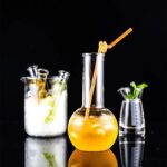 Beaker with yellow liquid and straw, laboratory glassware with scientific liquids, and a small plant in a beaker on a black background.