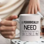 White ceramic mug with humorous text "I Periodically Need" and a periodic table of elements, perfect for science enthusiasts and coffee lovers.