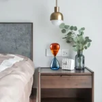 A modern bedroom nightstand with a colorful hourglass, a white clock, and a vase with eucalyptus branches, illuminated by a stylish hanging lamp.