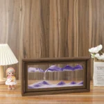 Small mountain landscape diorama with layered purple and white mountains inside a wooden frame, displayed on a wooden surface with decorative items around.