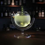 Glass fishbowl with a small plant floating inside, illuminated with a soft glow, set against a dark background with shelves of bottles.