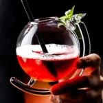 A vibrant red cocktail served in a clear glass teapot, garnished with fresh herbs and two black straws, held by a person's hand against a dark background.