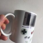 White ceramic mug designed to look like a classic Nintendo Game Boy, featuring a screen, buttons, and D-pad, perfect for gaming enthusiasts and retro collectors.