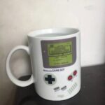 White ceramic mug designed to look like a classic Nintendo Game Boy, featuring a screen displaying "Super Mario Land" and colorful buttons, perfect for gaming enthusiasts.