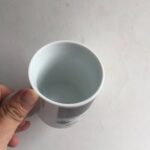 Empty white ceramic mug held by hand against a plain wall background.