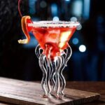 Jellyfish-shaped cocktail glass with a red drink and salt rim, featuring tentacle-like glass legs, on a wooden surface with a blurred dark background.