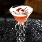 A stylish pink cocktail served in a unique glass with a tentacle-shaped stand, garnished with rose petals and herbs, perfect for a sophisticated party or special occasion.