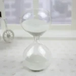Clear glass sand timer with white sand, placed on a white surface in a bright room, illustrating a classic time-measuring device for science and educational purposes.