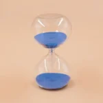 An image of a classic hourglass timer filled with vibrant blue sand, set against a neutral background. Perfect for educational demonstrations and science decor.