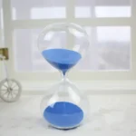 Clear glass sand timer with blue sand, measuring time visually, placed on a white surface near a window.