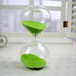 Transparent hourglass with green sand flowing from top to bottom, demonstrating the passage of time, placed on a white surface with a blurred background.
