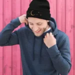 Young man wearing a black beanie with moon and star designs, smiling and adjusting his hat outdoors.