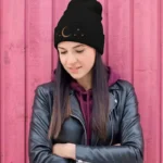 Young woman wearing a black beanie with moon and star design, black leather jacket, and purple hoodie, standing against a pink wooden background.