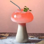 Mushroom-shaped beverage with pink foam, mint garnish, and metal straw on a pink background.