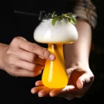 A colorful science beaker filled with yellow liquid, topped with foam, garnished with herbs, and held by a person's hands, showcasing a creative science experiment.