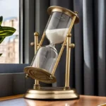 Elegant sand timer with a gold frame and glass body, filled with white sand, placed on a wooden surface near a window.