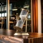 Brass and glass hourglass timer with flowing sand, placed on a wooden table in a contemporary interior space, showcasing a stylish and functional decorative piece.