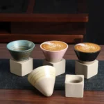 A set of three ceramic coffee cups with latte art, each placed on a minimalist concrete stand, accompanied by an additional cone-shaped cup and a small square holder, perfect for modern decor.