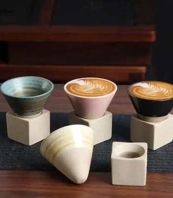 A set of three ceramic coffee cups with latte art, each placed on a minimalist concrete stand, accompanied by an additional cone-shaped cup and a small square holder, perfect for modern decor.
