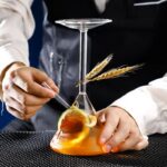 Close-up of a scientist's hands conducting a chemistry experiment with a flask containing amber liquid, garnished with wheat stalks, in a laboratory setting.