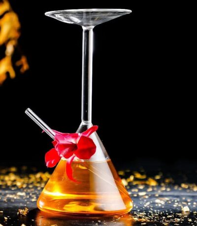 Erlenmeyer flask with a glass stirring rod and a glass dish balanced on top, filled with a yellow-orange liquid, set against a black background with scattered gold particles.
