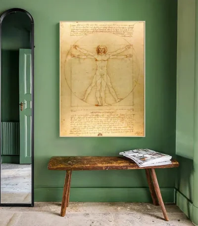 Classic Vitruvian Man drawing by Leonardo da Vinci displayed on a green wall, with a rustic wooden bench and a tall mirror in a well-lit room.