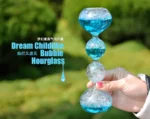 Hand holding a colorful, transparent bubble hourglass with blue and clear sections, set outdoors with a blurred green background.