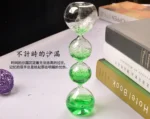 Glass hourglass with green liquid and bubbles, placed on a white surface with books in the background.