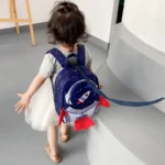 Adorable child with a navy blue rocket backpack, walking indoors near a curved white wall, dressed in a grey t-shirt and white skirt, engaging in playful learning.