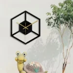 Stylish modern wall clock with a black hexagonal frame, featuring gold clock hands, mounted on a white wall next to a potted plant and a playful frog figurine.