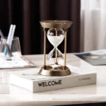 Elegant brass sand timer with white sand, placed on a white book titled "WELCOME" in a modern workspace setting.