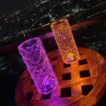 Color-changing LED glasses with textured surface, illuminated in purple and orange, on a wooden table overlooking a cityscape at night.